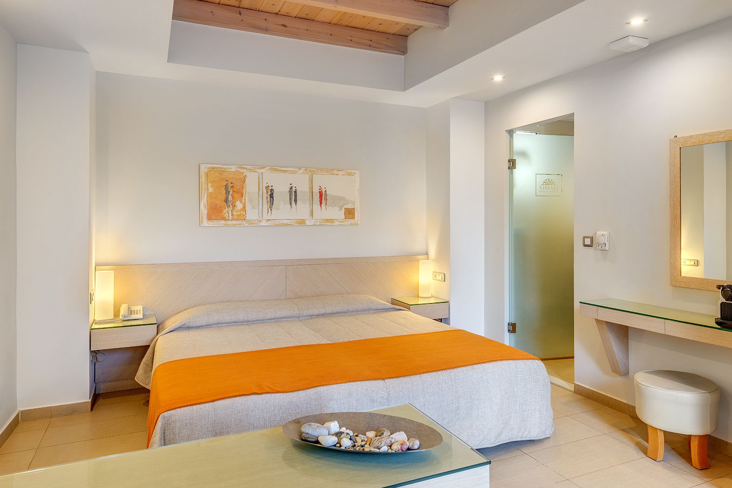 A hotel room with a large bed, bedside lamps, a glass table with a bowl of stones, and a mirror above the sink.