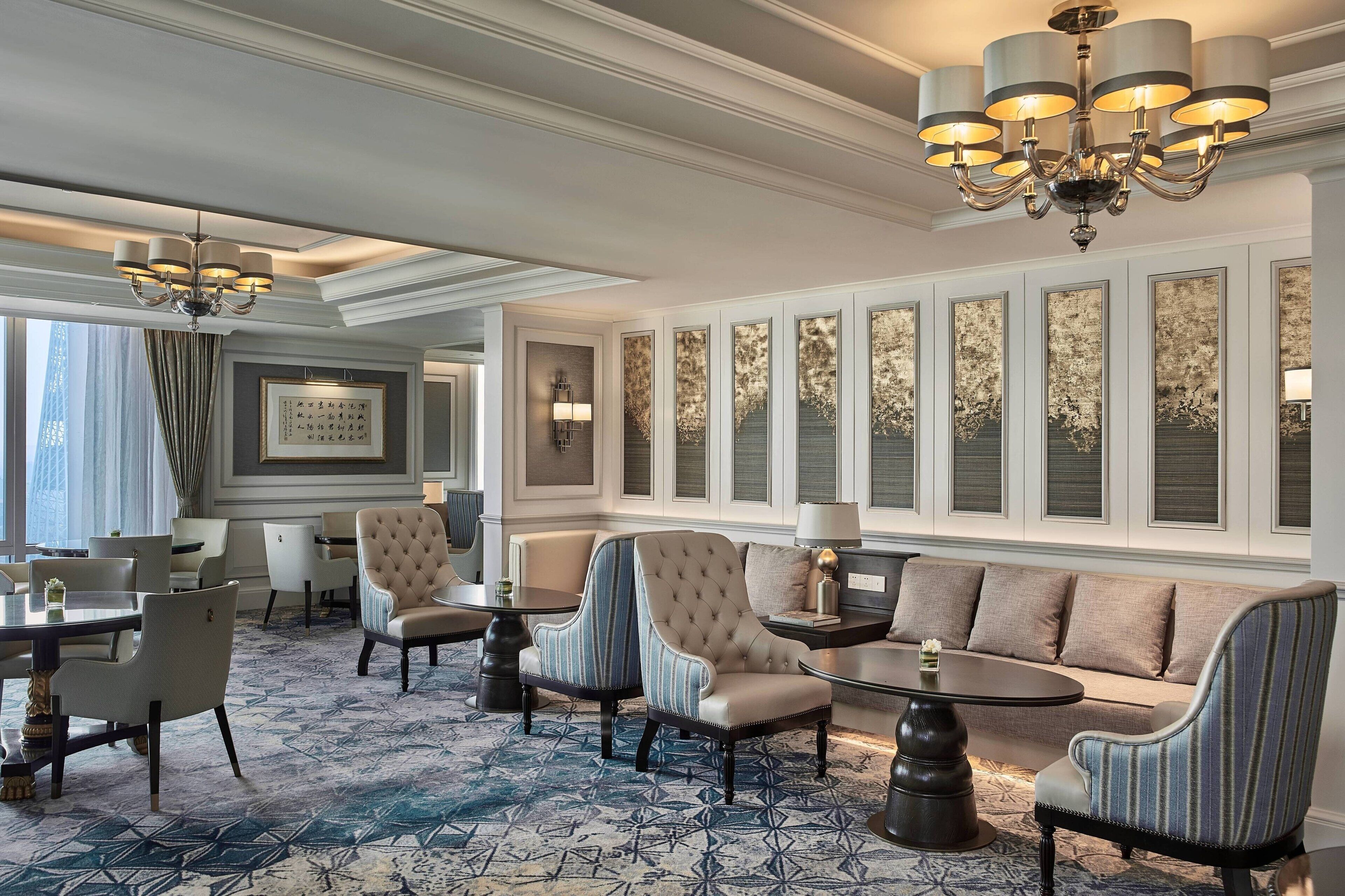 A hotel lobby with a patterned carpet, a sofa, armchairs, a chandelier, and framed artwork on the wall.