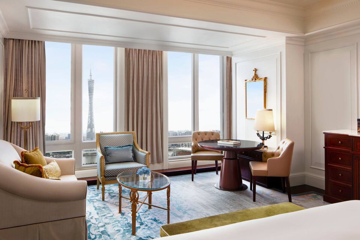 A hotel room with a large window, a sofa, a chair, a table, a lamp, and a dresser.