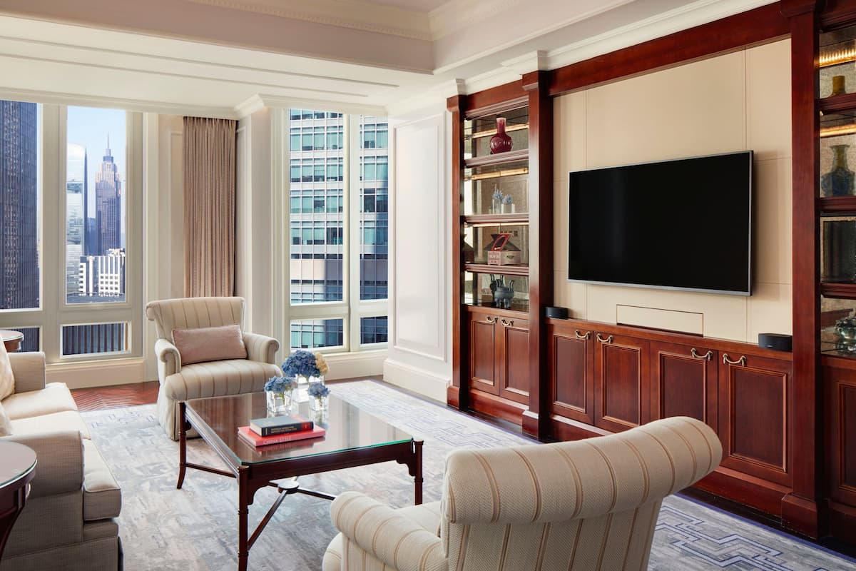 A spacious living room with a large window offering a city view, a flat-screen TV mounted on a wooden panel, and a comfortable seating area with a coffee table.