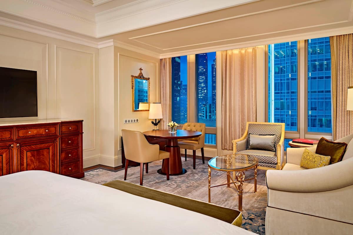 A hotel room with a large bed, a wooden dresser, a dining table with chairs, a sofa, and a large window with a city view.