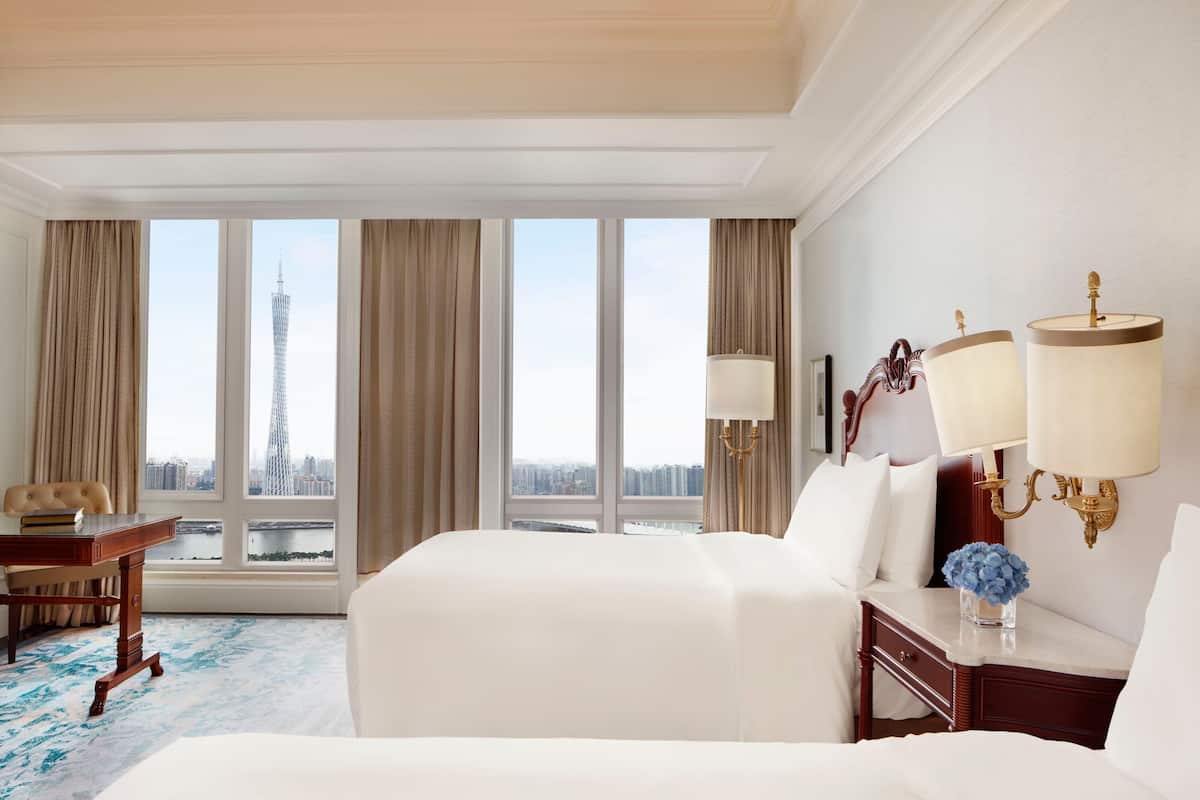 A hotel room with a large bed, a desk, a chair, a lamp, and a view of a tall building through the windows.