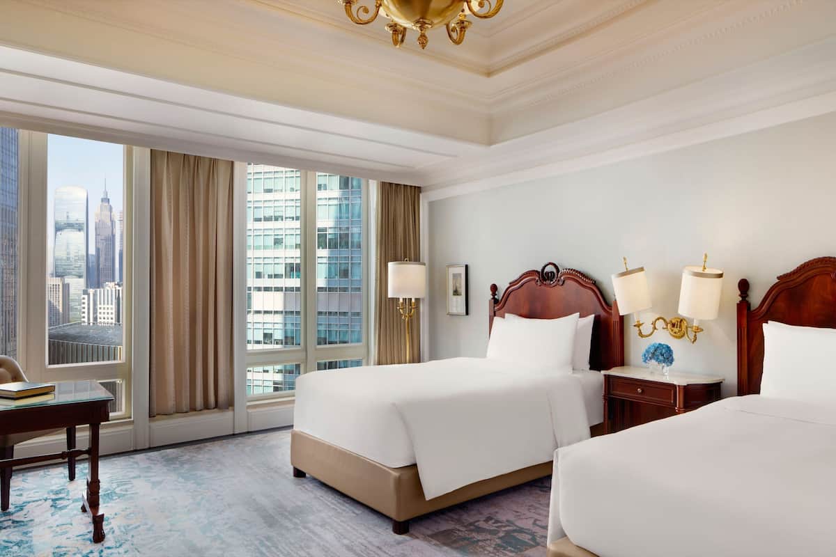 A hotel room with two beds, a desk, and a view of the cityscape.