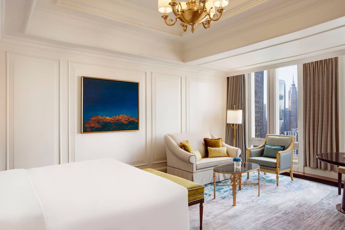 A hotel room with a large bed, a sofa, a chair, a small table, and a painting on the wall.