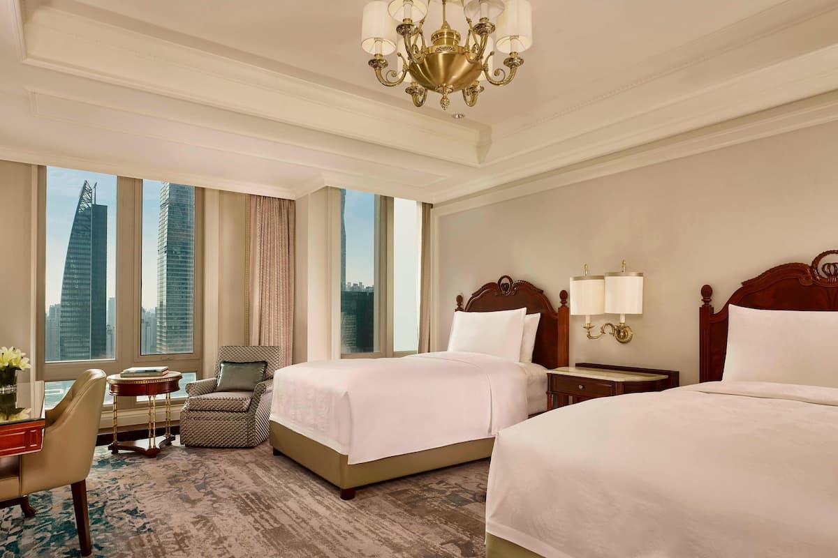 A hotel room with two beds, a desk with a chair, a small table, and a view of tall buildings.