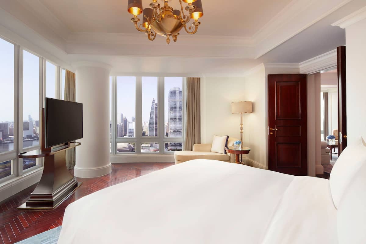 A hotel room with a large bed, a television on a stand, a sofa, a lamp, and a view of the cityscape through large windows.