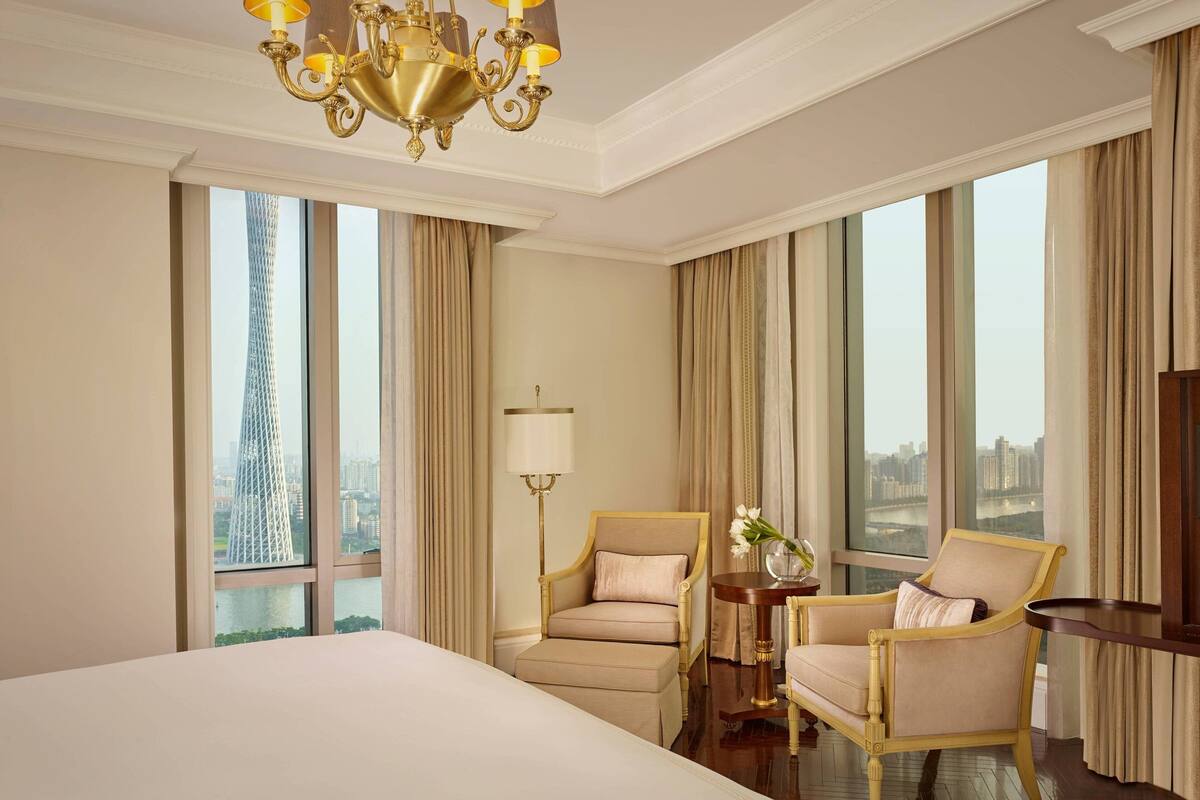 A hotel room with a large bed, two armchairs, a side table with a vase, and a chandelier.