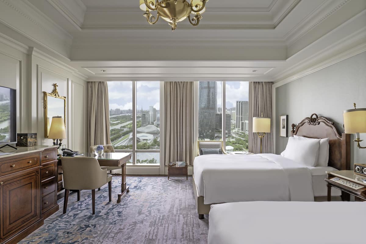 A hotel room with two beds, a desk, a chair, a TV, and a large window with a city view.