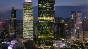 A cityscape at night with illuminated skyscrapers, a curved building with a green-lit facade, and a modern architectural structure with a unique roof design.