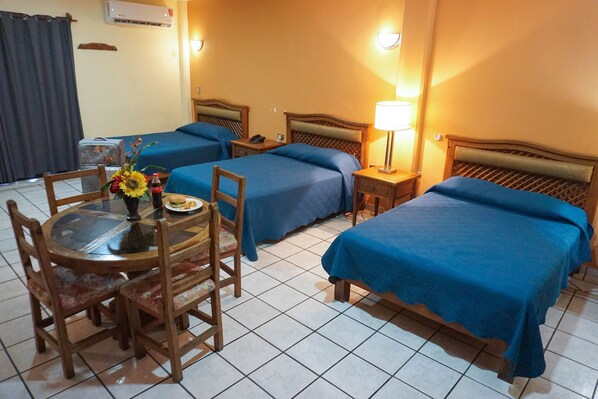 Habitación de hotel con dos camas, una mesa de comedor con sillas, una mesita con un florero y aperitivos, y un aire acondicionado instalado en la pared.