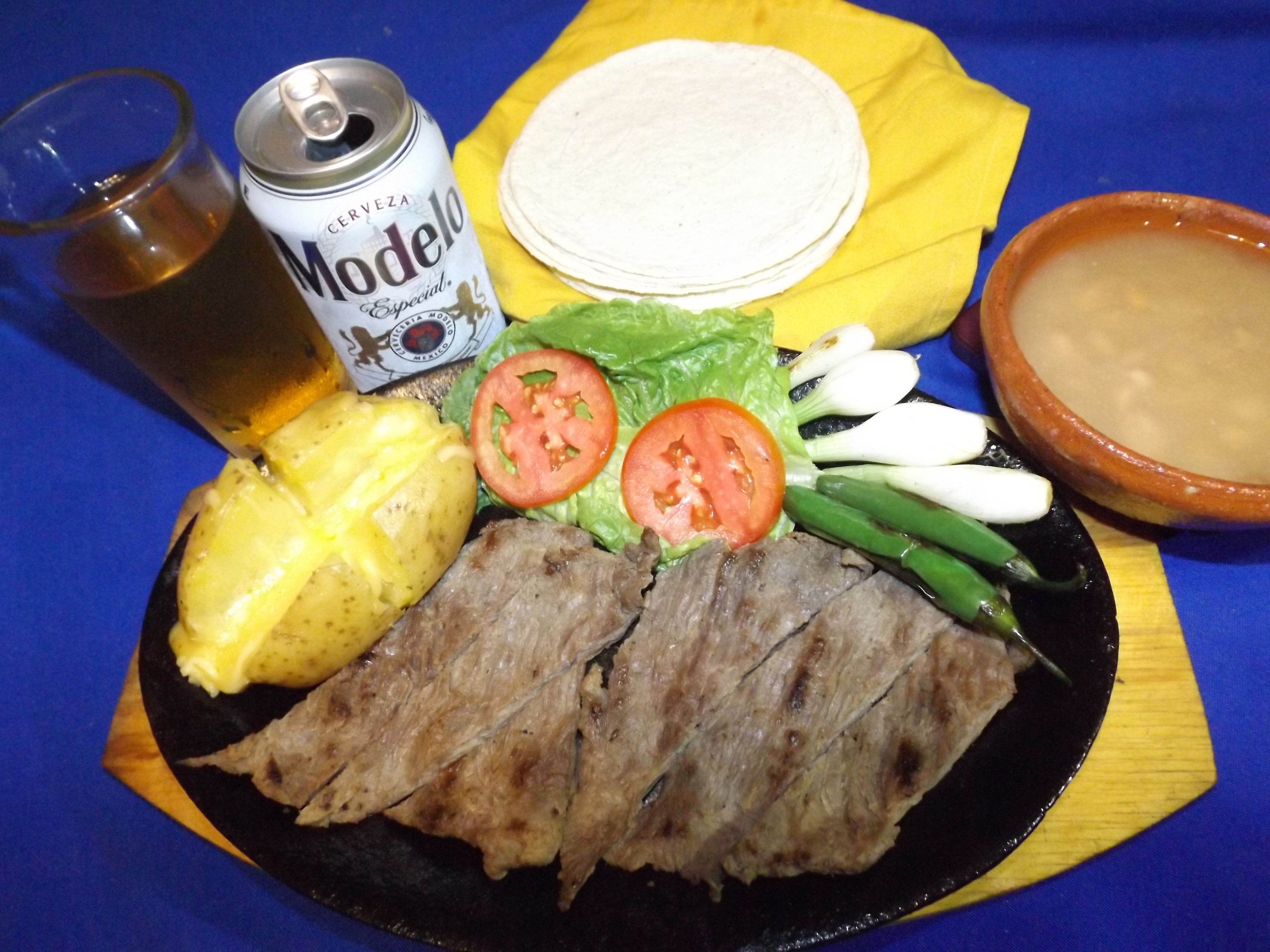Une assiette contenant de la viande grillée, des pommes de terre, de la laitue, des tomates et des oignons verts, une canette de bière Modelo, un verre de bière et un bol de soupe, le tout posé sur une nappe bleue.