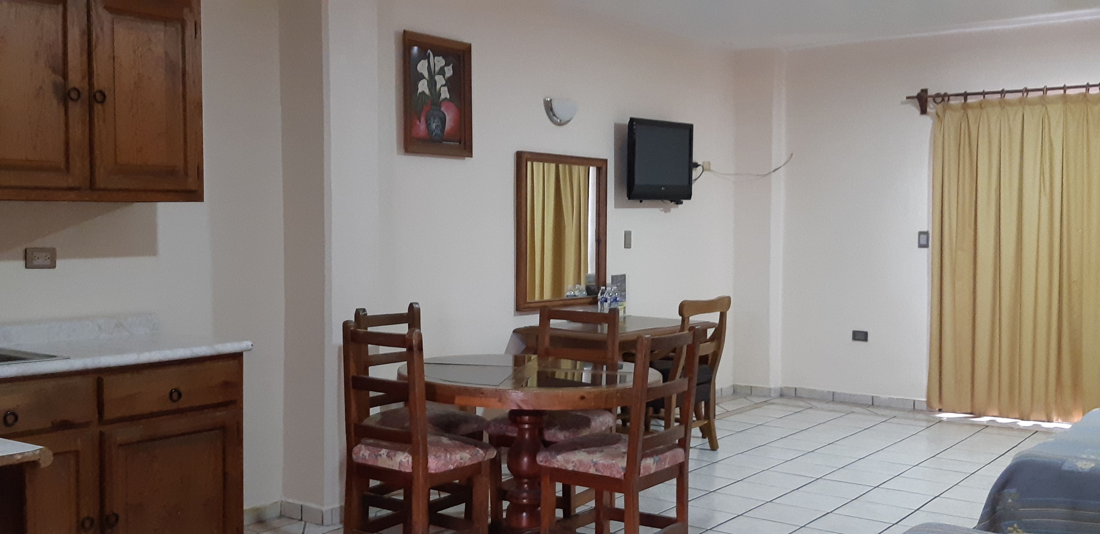 A dining area with a wooden table and chairs, a television mounted on the wall, and a window with curtains.