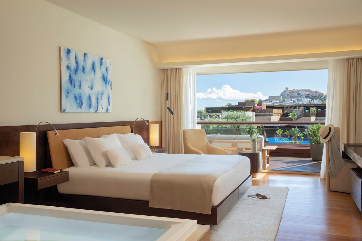 Ein Hotelzimmer mit einem großen Bett, einem Schreibtisch, einem Stuhl, Blick auf eine Stadtlandschaft und einem Balkon mit Pflanzen.