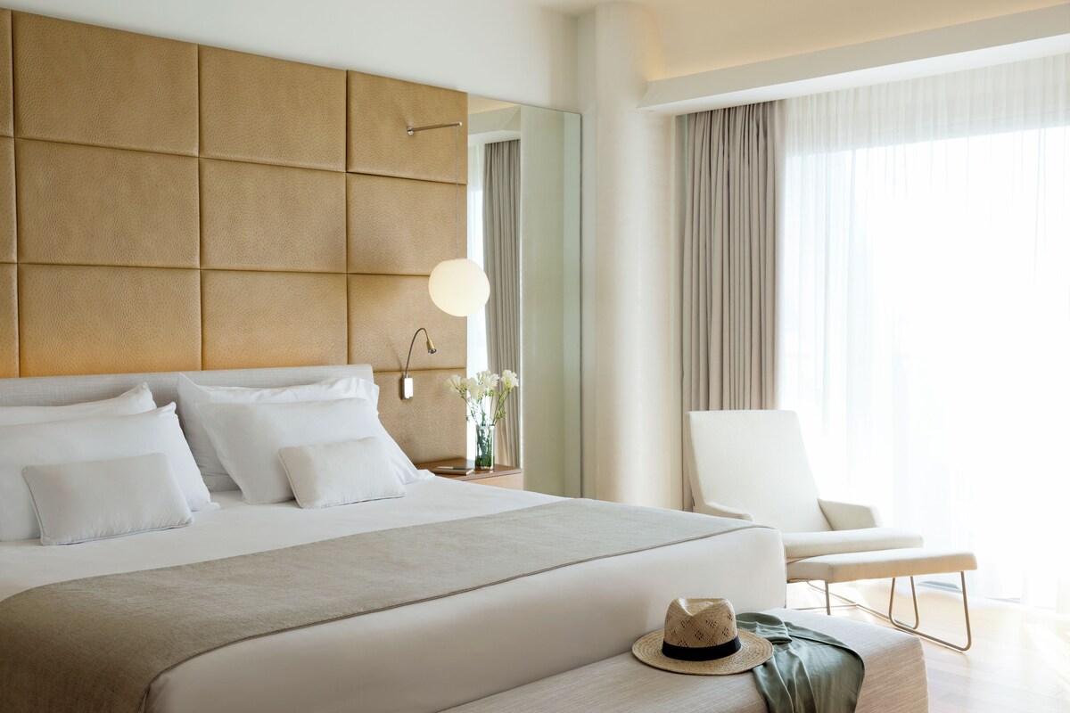 A modern hotel room with a large bed, a headboard with a geometric pattern, a bedside table with a lamp, a chair, and a window with curtains.