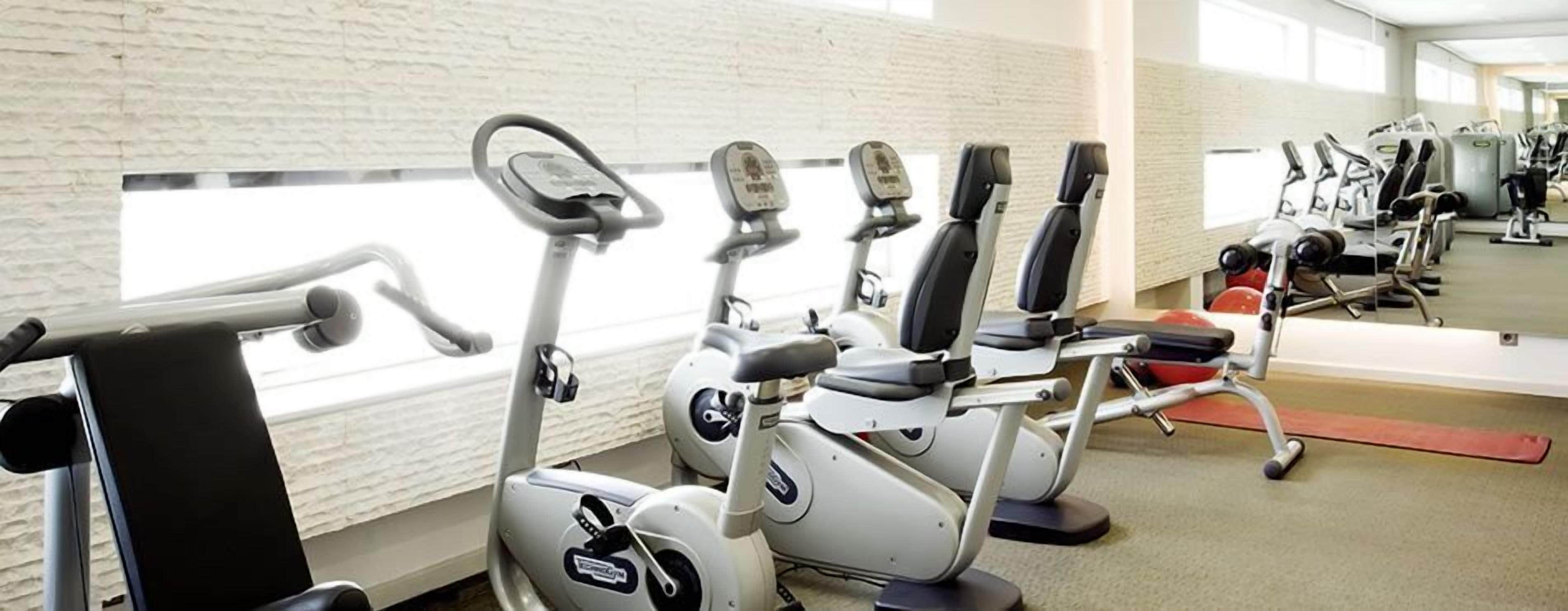 A gym with stationary bikes and elliptical machines, a mirror on the wall, and a person using a rowing machine.