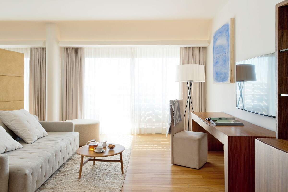 Ein modernes Hotelzimmer mit einer Couch, einem kleinen runden Tisch mit Teekanne, einem Schreibtisch mit Lampe und einem großen Fenster mit Vorhängen.