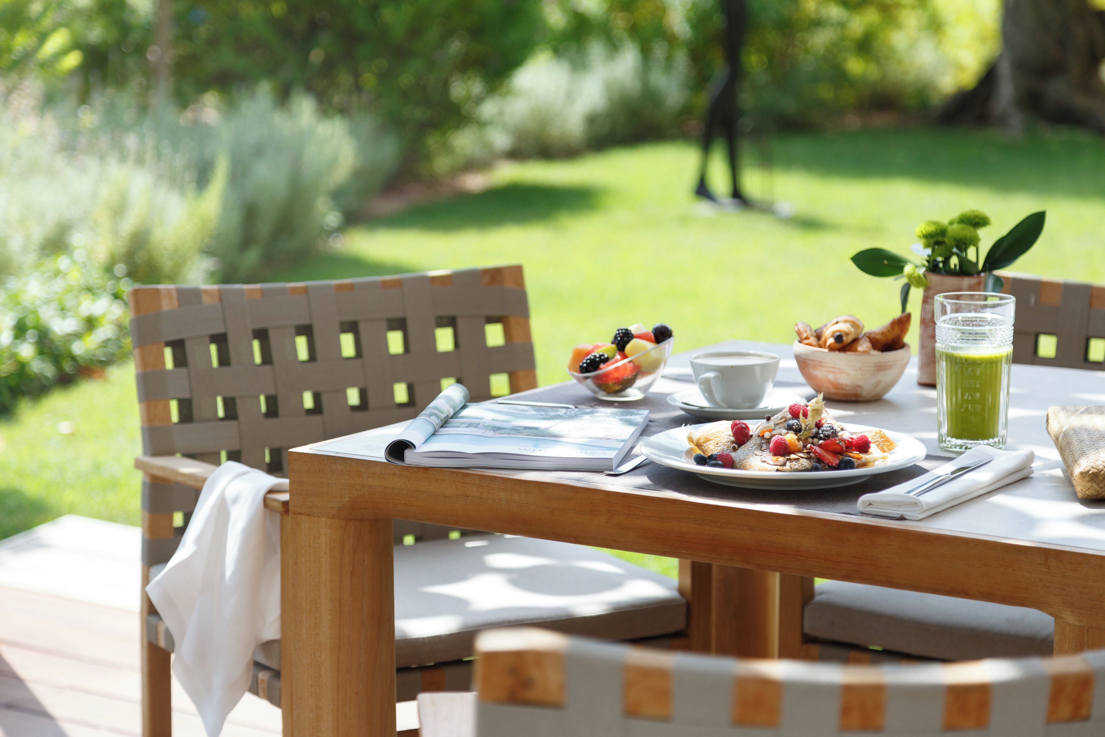 Una mesa de madera con un desayuno que incluye un bol de fruta, un plato de cereales, una taza y un vaso de jugo verde.