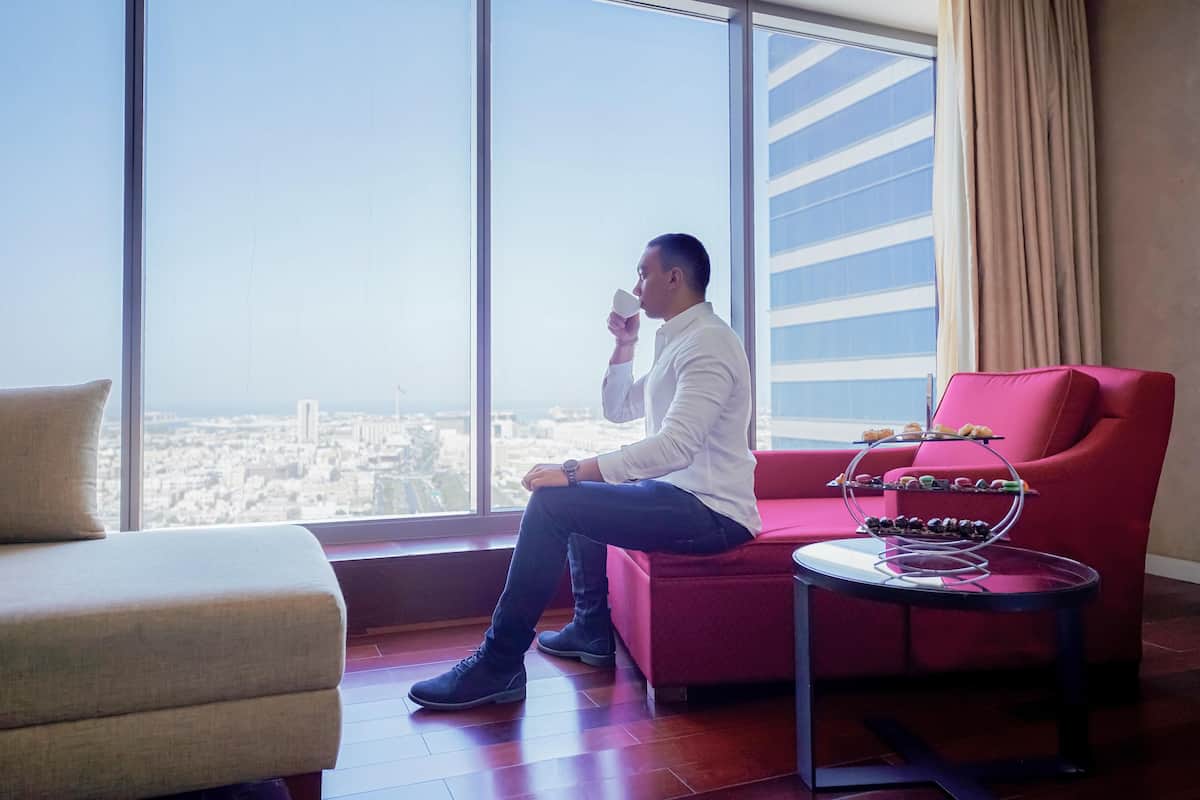 A man sitting on a red sofa by a large window with a city view, a small table with a glass top, and a beige armchair.