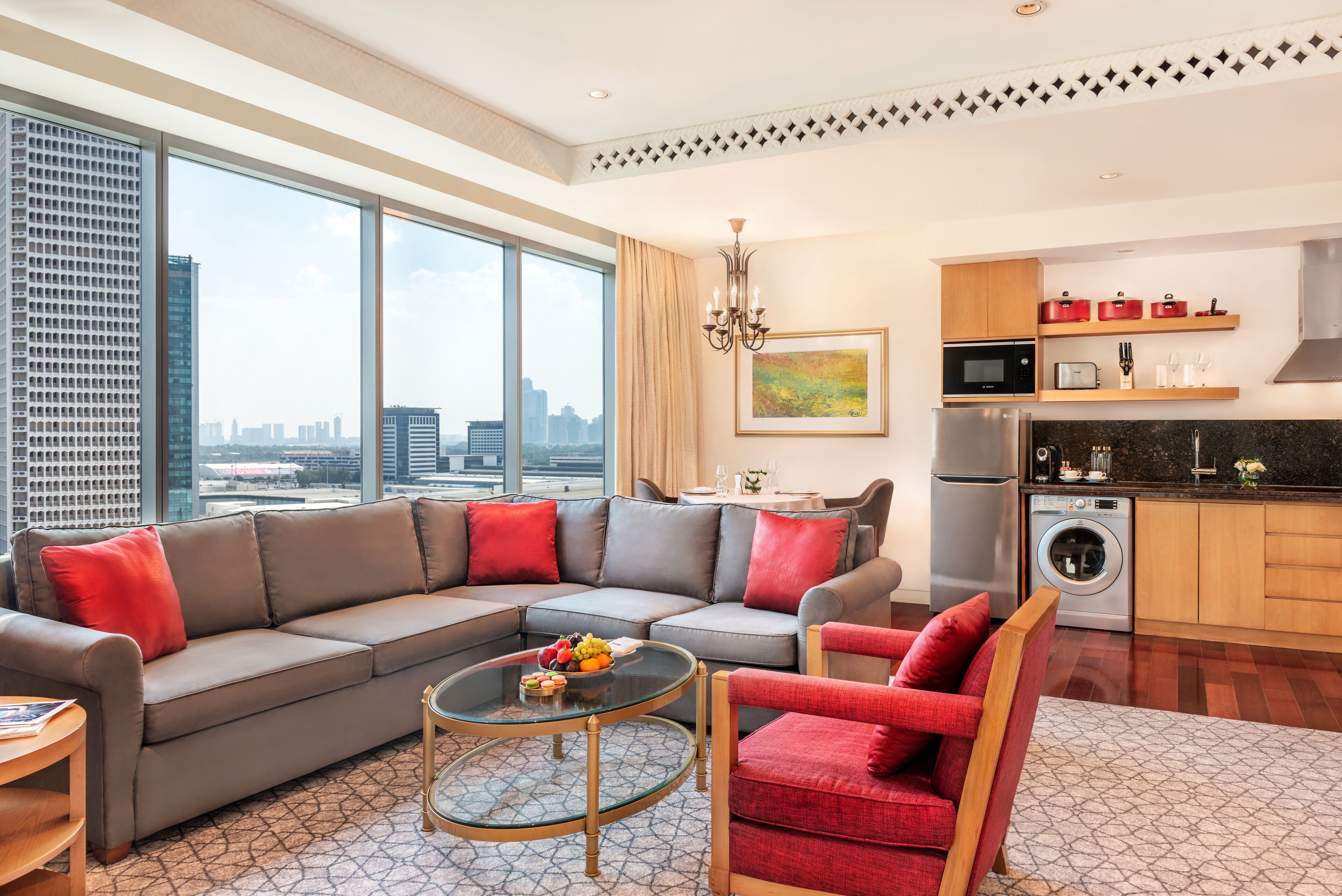 A modern living room with a grey sectional sofa, red armchairs, a glass coffee table, and a city view through large windows.