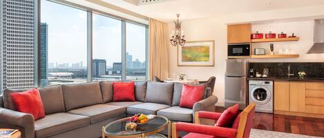 A modern living room with a grey sectional sofa, red armchairs, a glass coffee table, and a city view through large windows.