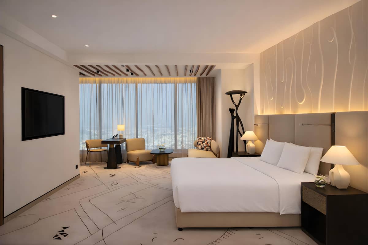 A hotel room with a large bed, a TV, a desk with a chair, a lamp, and a view of the city through large windows.