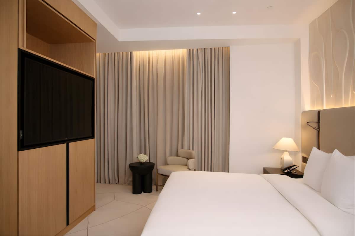 A modern hotel room with a large bed, a flat-screen TV, a seating area with a chair and a small table, and a nightstand with a lamp and phone.