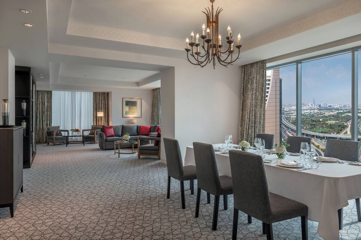 A hotel room with a dining area, a sofa set, and a large window offering a city view.