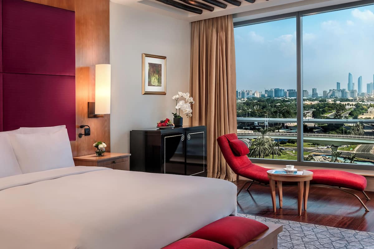 A hotel room with a large bed, a desk, a red chair, and a view of a cityscape through a large window.
