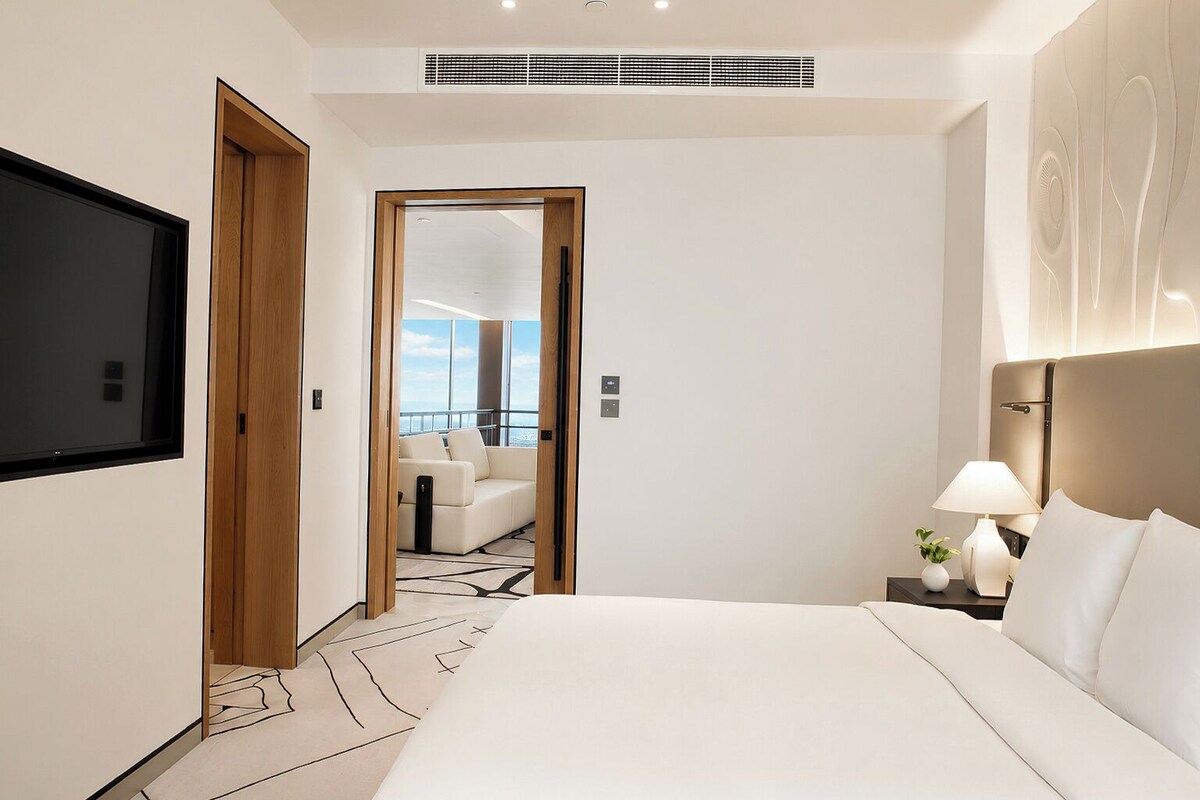 A modern hotel room with a large bed, a TV, a bedside table with a lamp, and a view of the city through a balcony door.