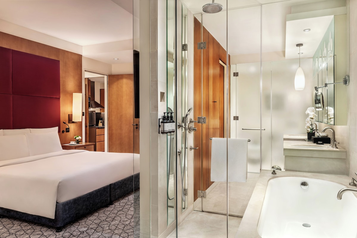 A modern hotel room with a large bed, a glass-enclosed shower, and a separate bathroom with a large bathtub and sink.