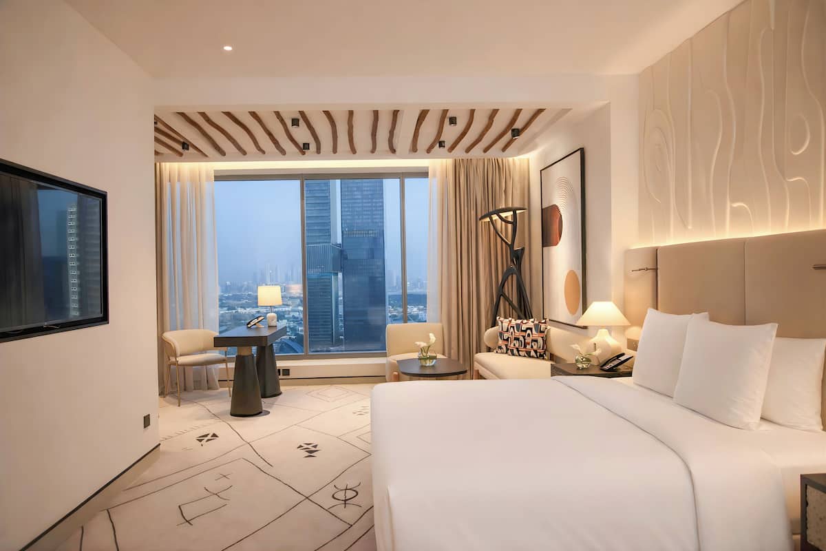 A modern hotel room with a large bed, a desk, a chair, and a view of the cityscape through large windows.