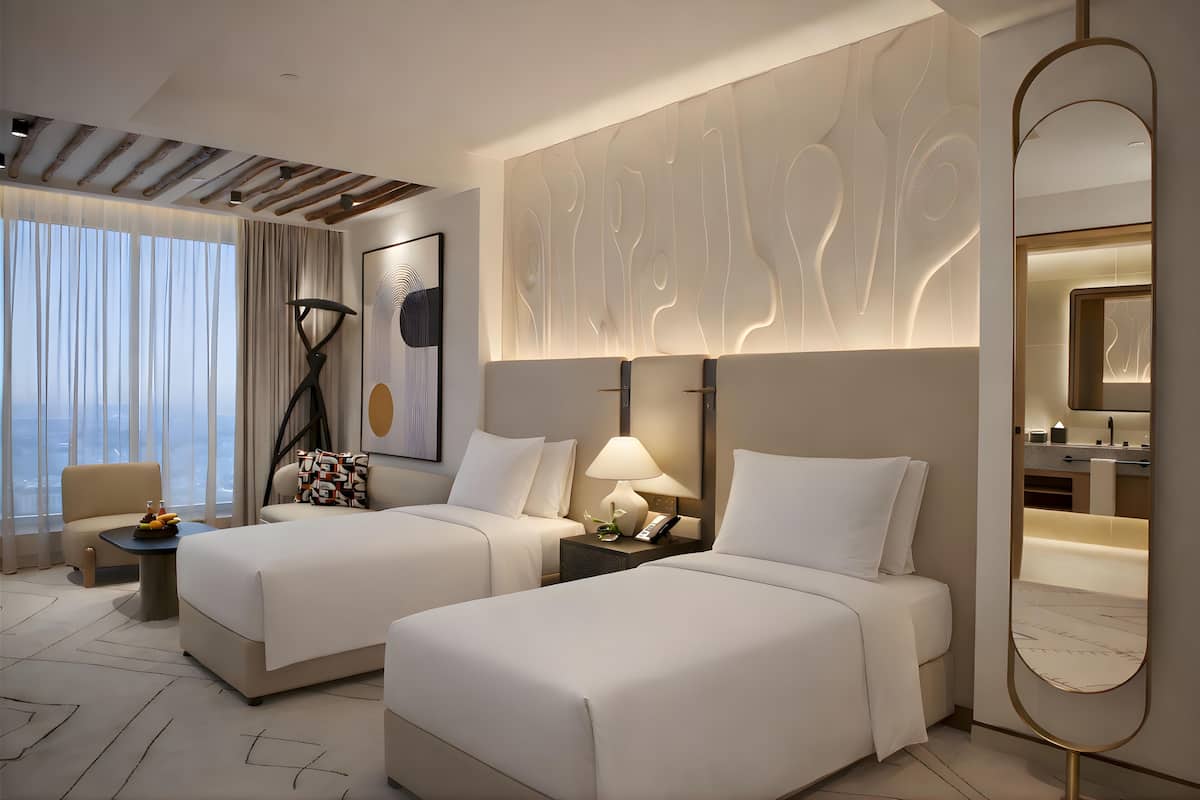A hotel room with two beds, a sitting area with a chair and a small table, a large mirror, and a view of the city through large windows.