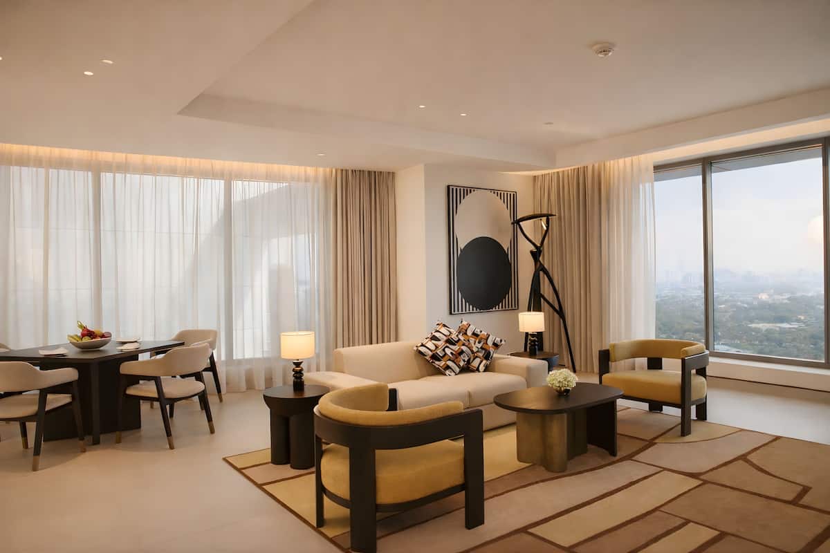 A modern hotel room with a dining area, a sofa set, a coffee table, and a large window offering a city view.