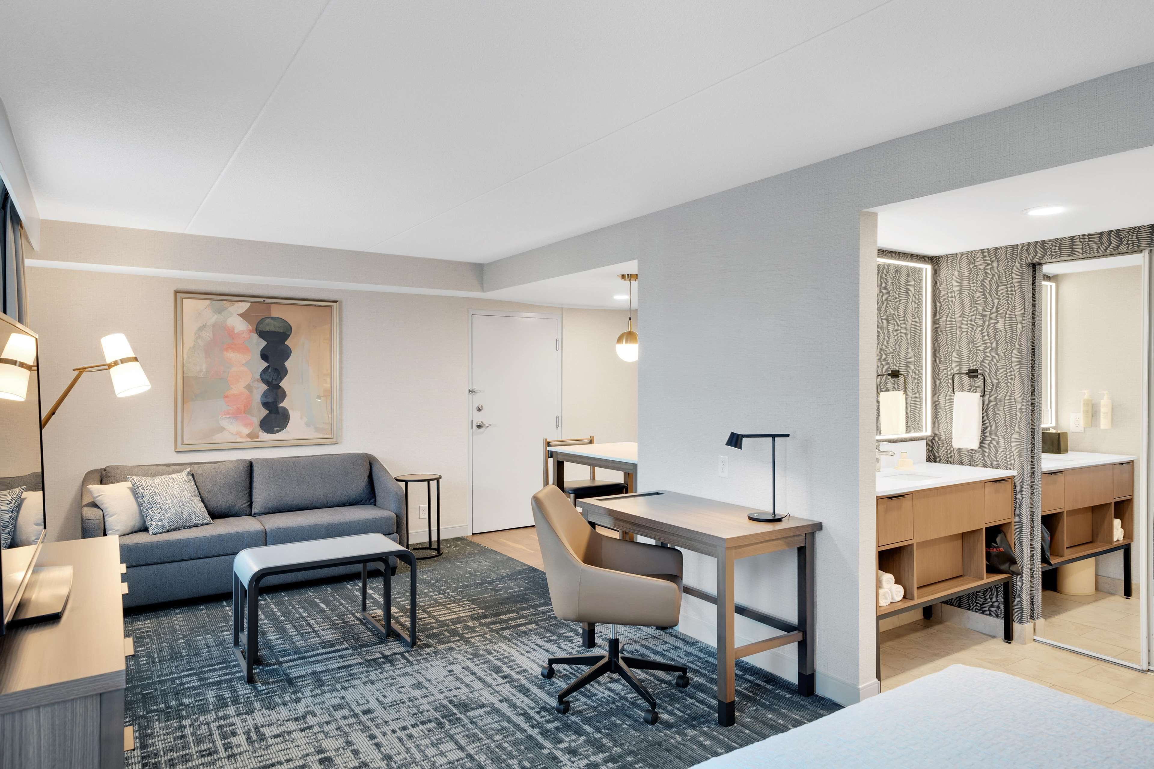 A modern hotel room with a grey sofa, a desk, a computer, a chair, a large mirror, and a bathroom with a vanity and sink.