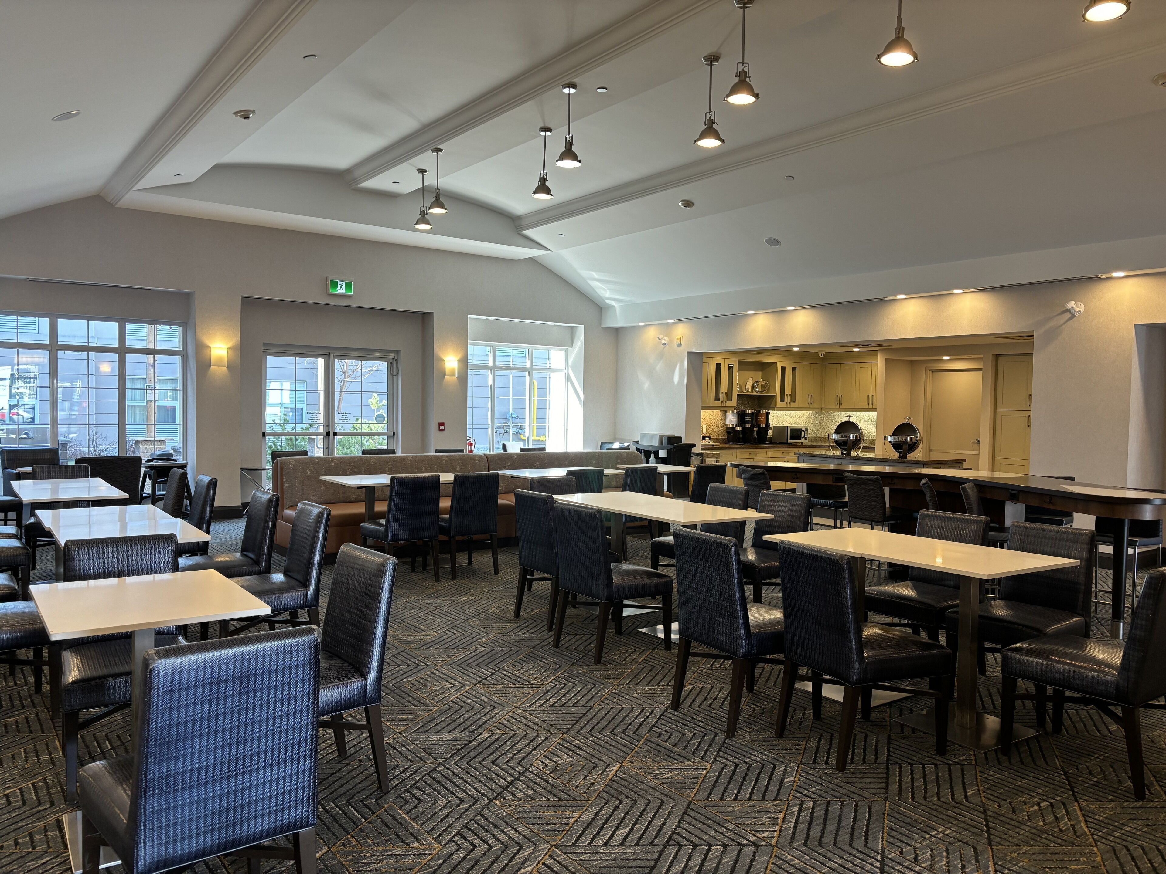 A restaurant with multiple tables and chairs, a kitchen area, and large windows allowing natural light.
