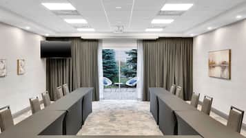Un salón de conferencias con una larga mesa, sillas, un televisor de pantalla plana y vistas a un área exterior con sillas a través de un vidrio.