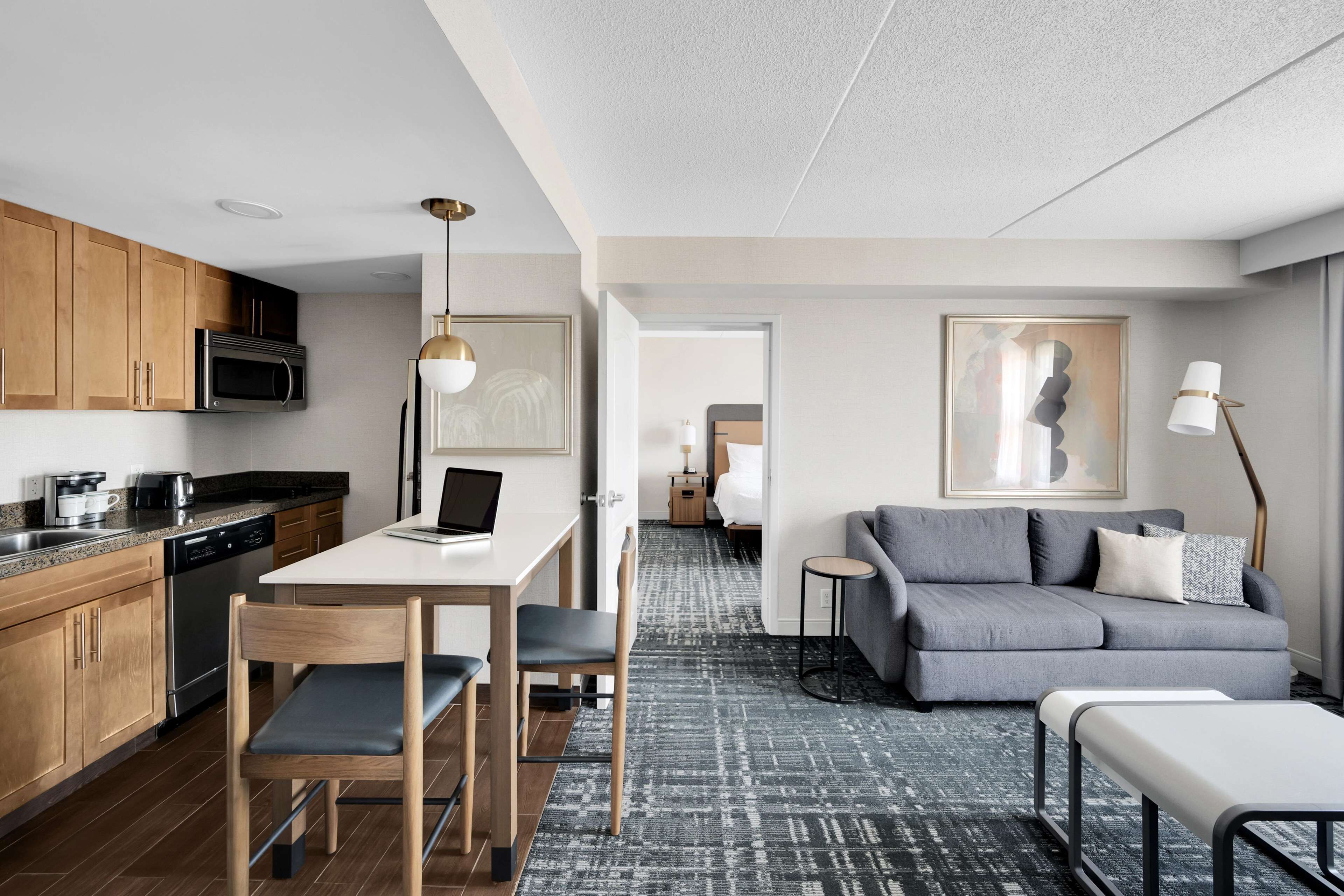 A modern kitchen with wooden cabinets, a microwave, and a dishwasher. There is a dining area with a table and chairs, and a grey sofa in the living room.
