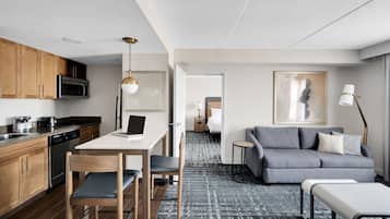 A modern kitchen with wooden cabinets, a microwave, and a dishwasher. There is a dining area with a table and chairs, and a grey sofa in the living room.