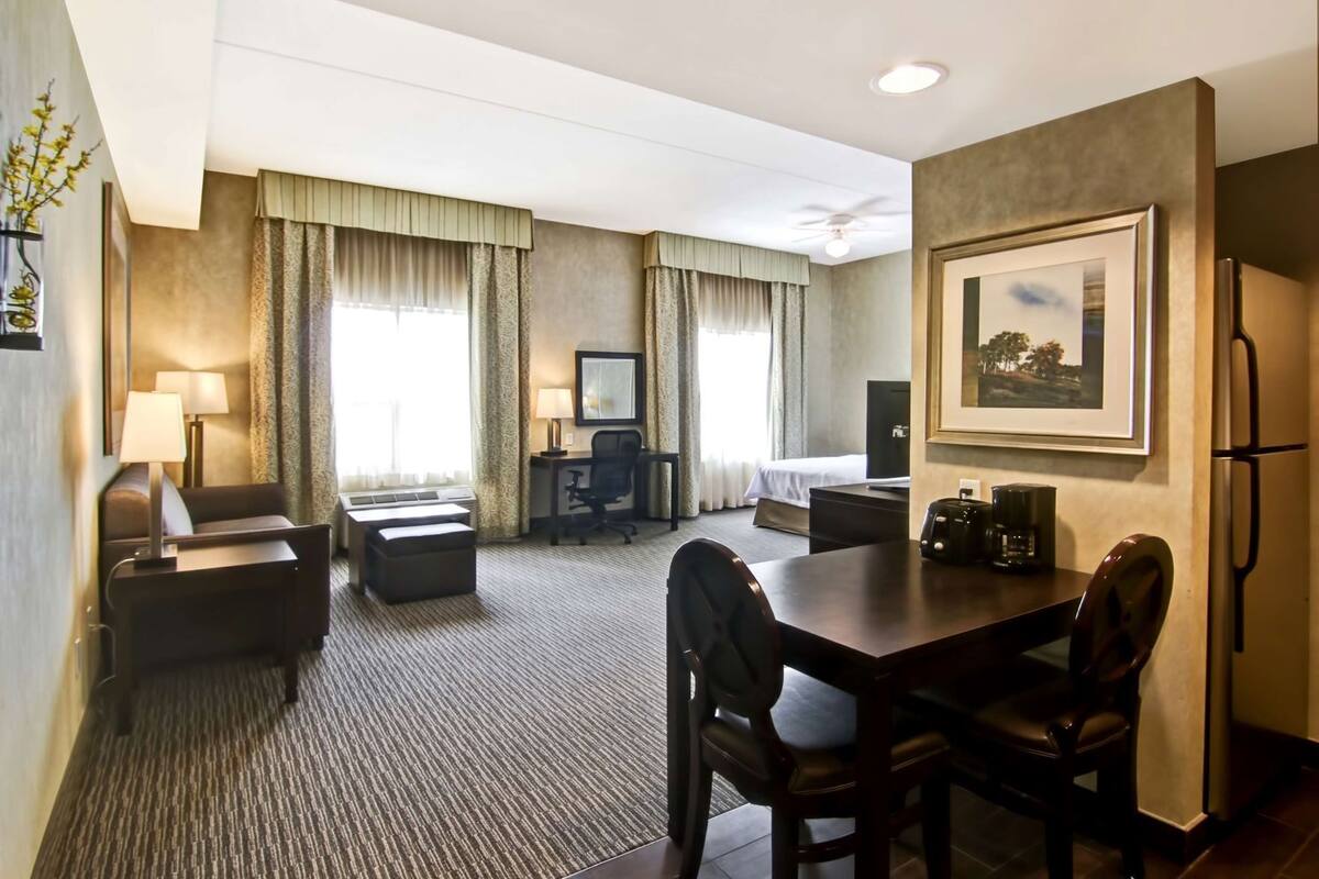 A hotel room with a bed, a desk, a chair, a sofa, a dining table, and a refrigerator.