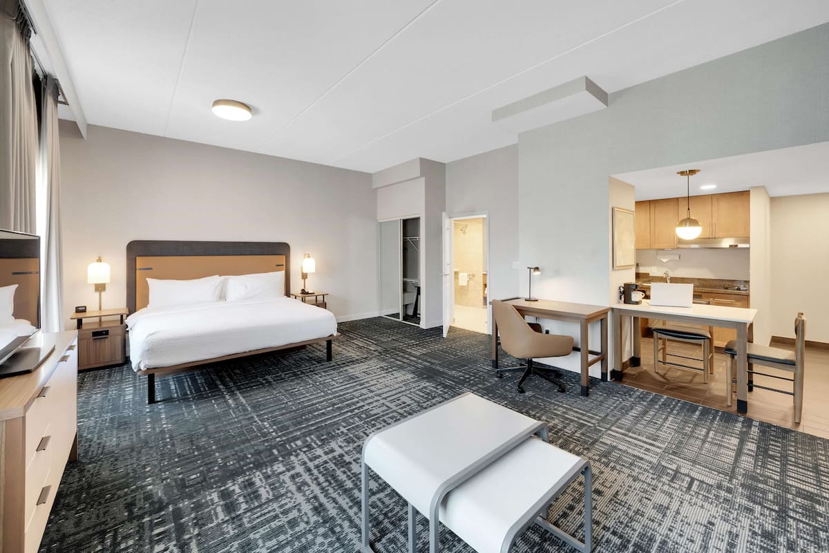 A hotel room with a large bed, a desk, a chair, a bench, a wardrobe, and a kitchenette.