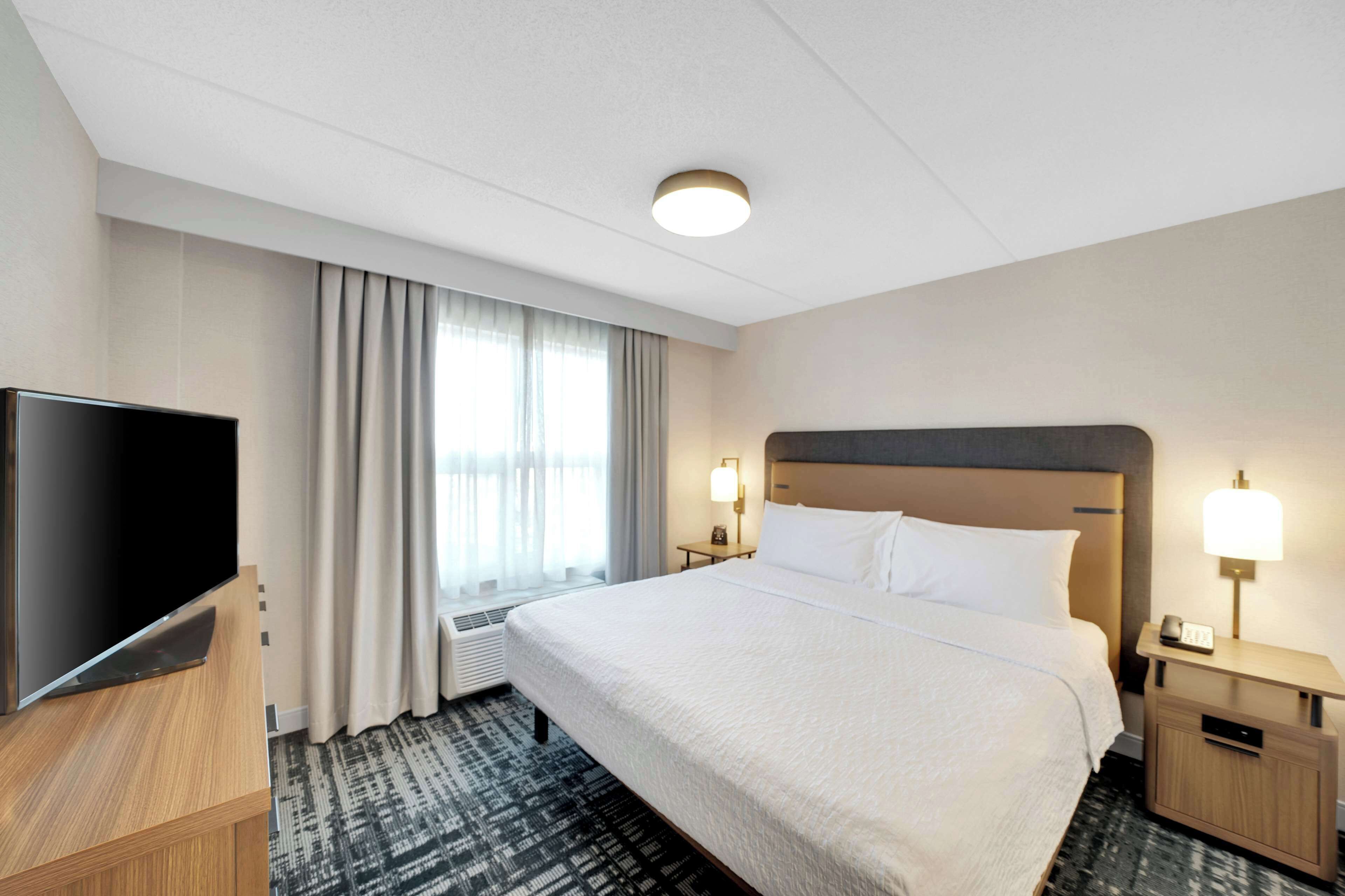 A hotel room with a large bed, a flat-screen TV, a bedside table with a lamp, and a window with curtains.