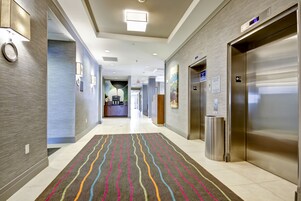 Corredor moderno de hotel com carpete estampado, elevadores e uma área de recepção ao fundo.
