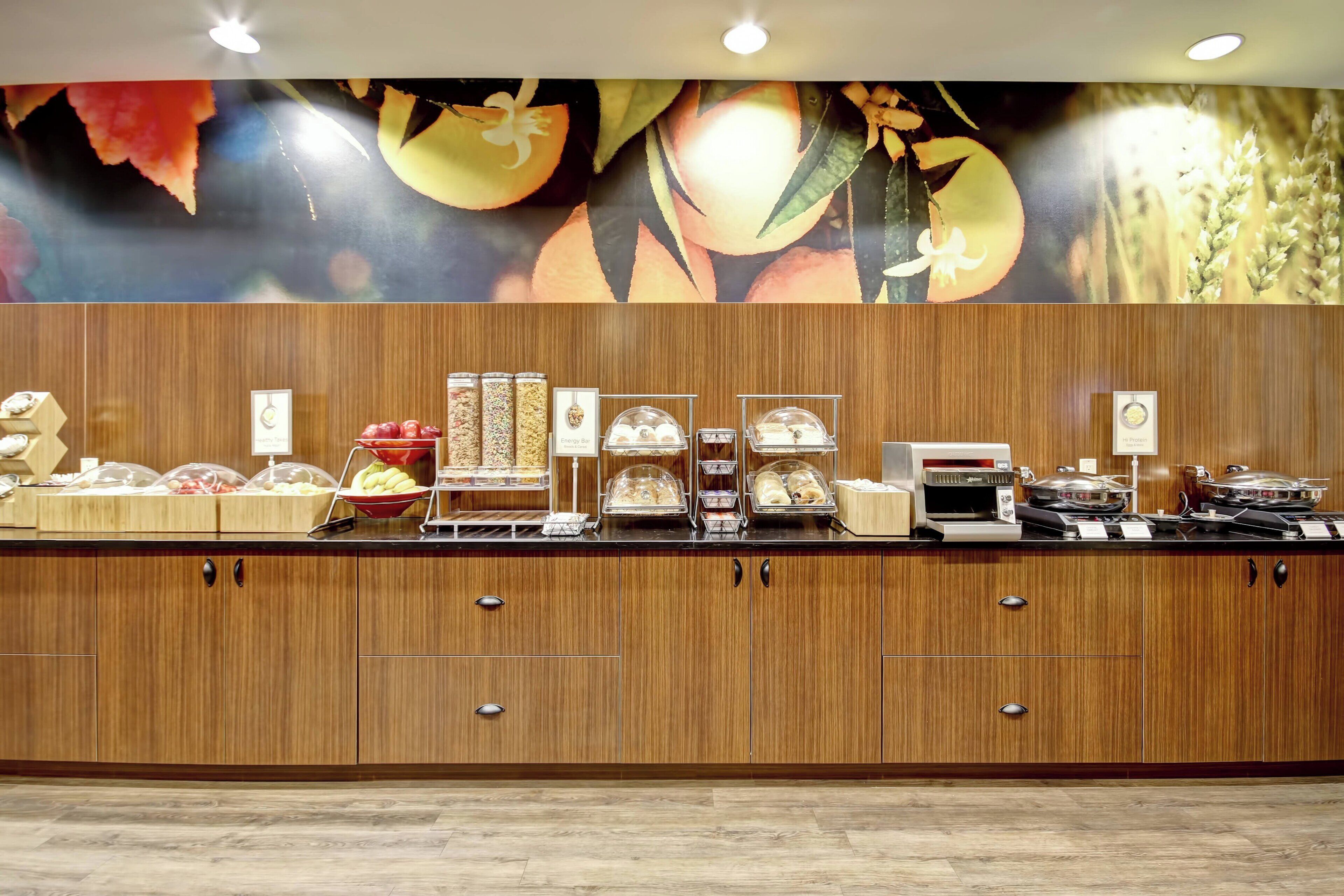 Mostrador de buffet con diferentes alimentos y una pared decorativa con motivos de frutas y flores.