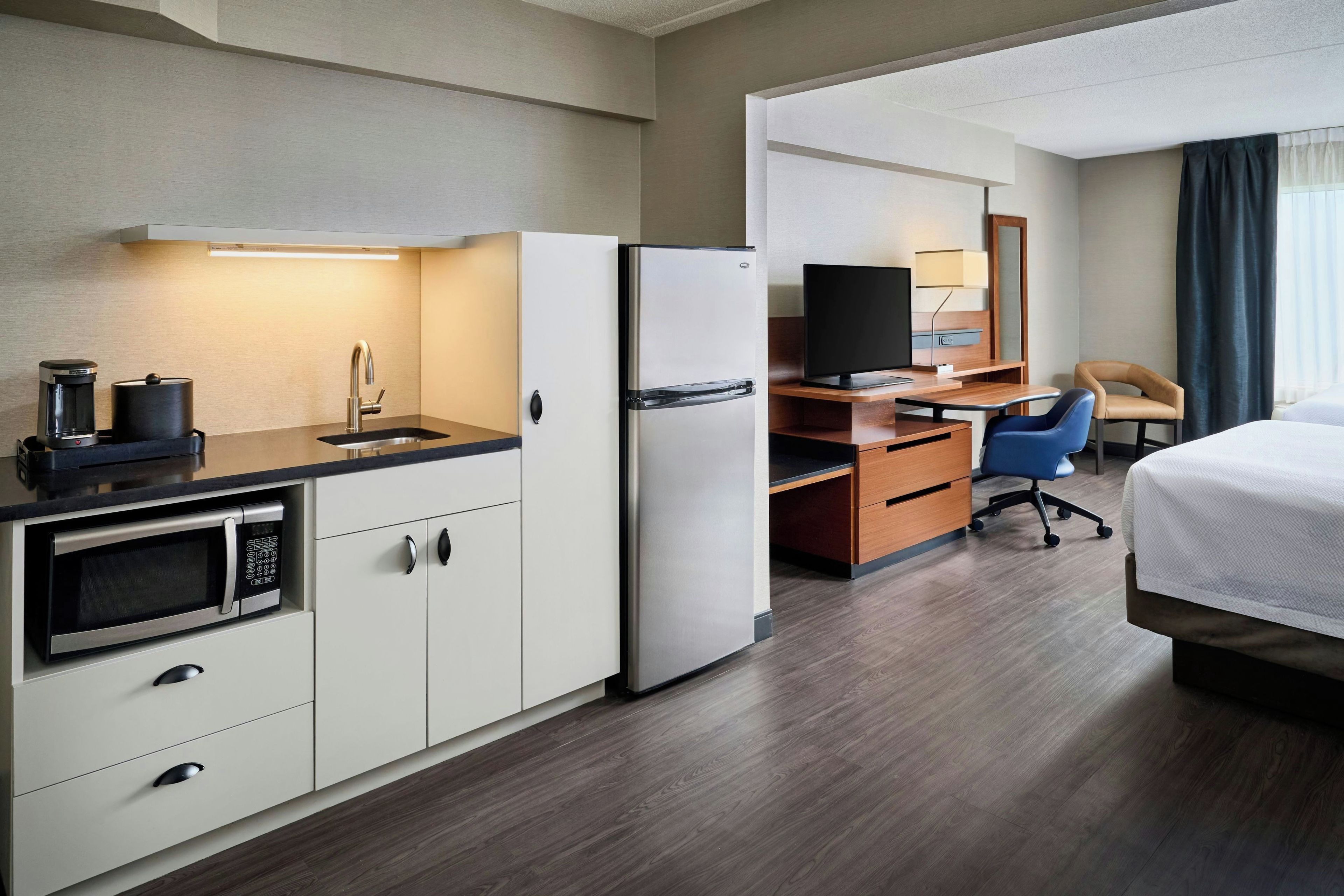 A hotel room with a kitchenette, a bed, a desk with a chair, and a television.