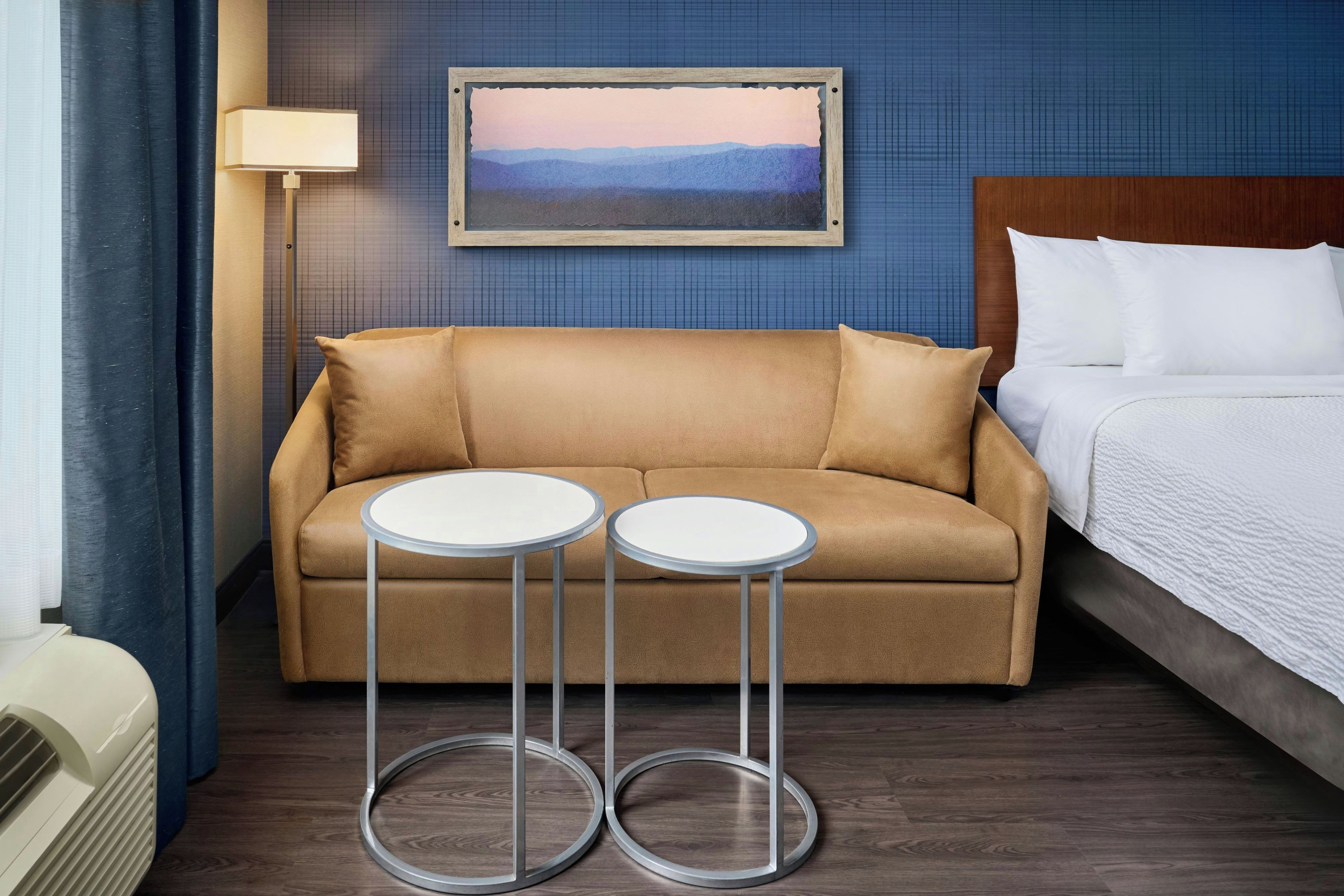 A hotel room with a sofa, two round tables, a bed, a painting on the wall, and a lamp.