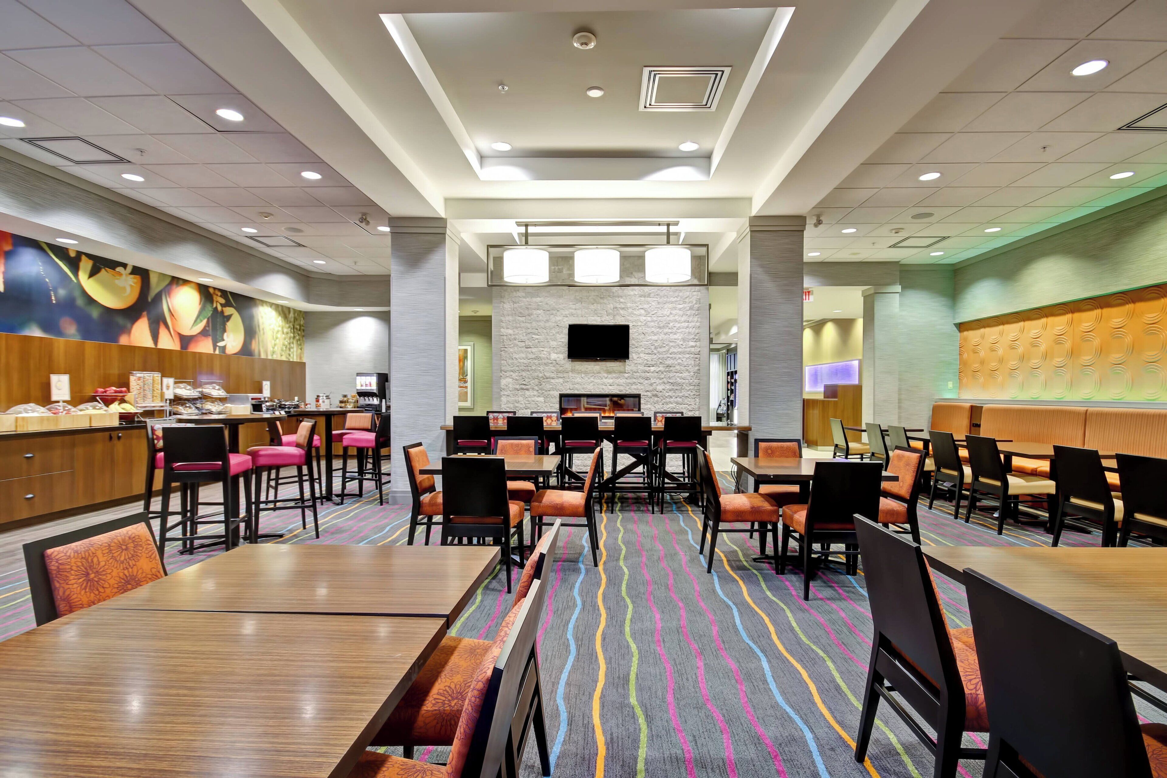 A hotel dining area with multiple tables and chairs, a central TV screen, and a colorful carpet.