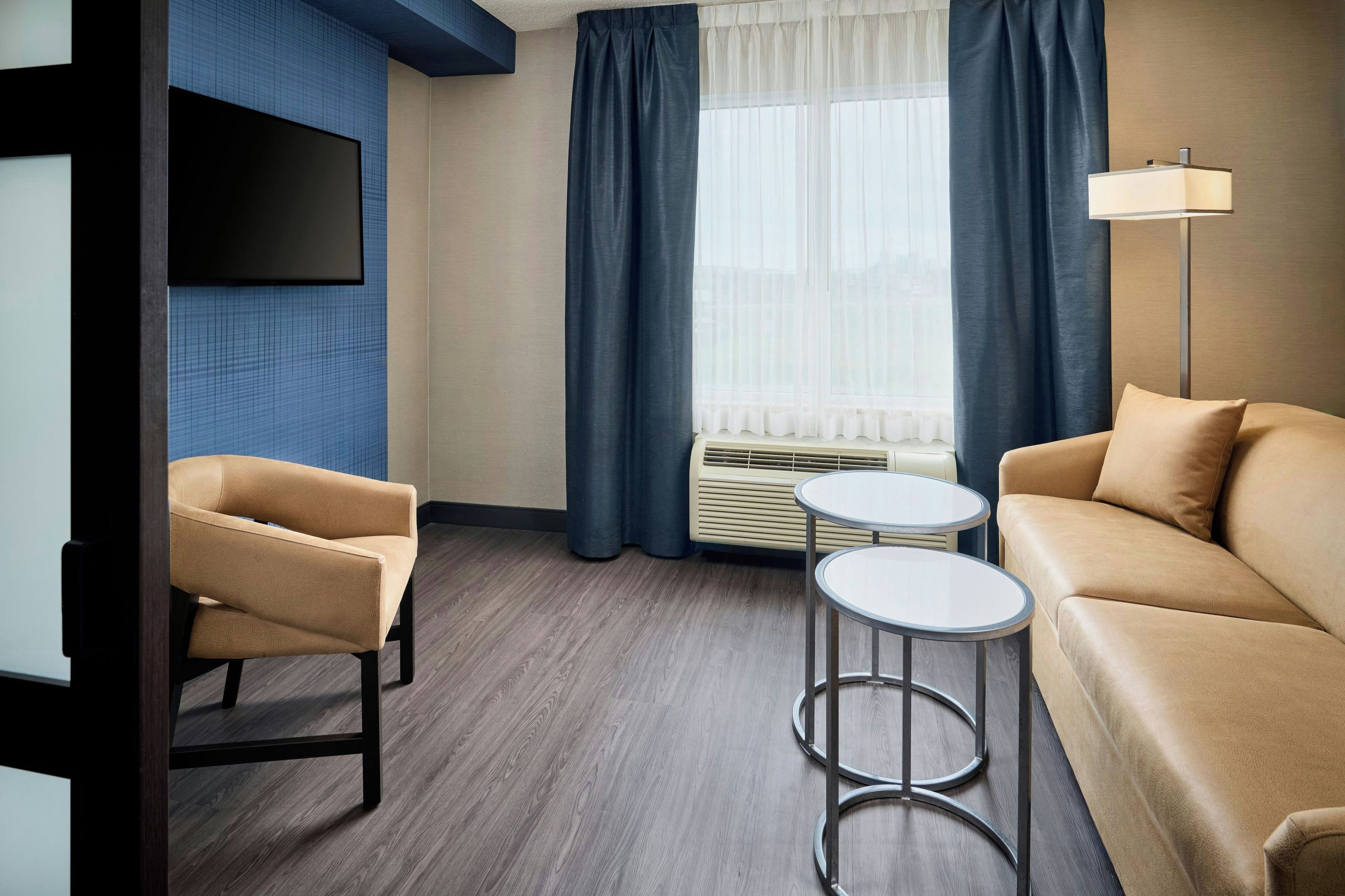 Una habitación de hotel moderna con sofá, dos sillones, una mesita, televisor montado en la pared y ventana con cortinas.