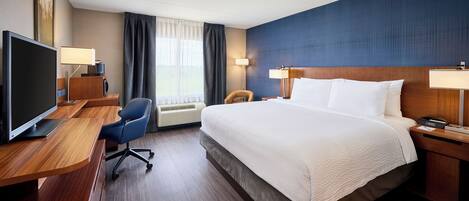 A hotel room with a bed, a desk with a TV, a chair, and a bedside table with a lamp.