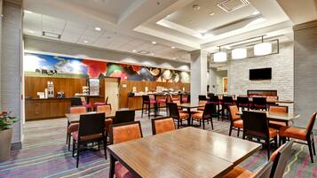 A hotel dining area with multiple tables and chairs, a kitchen area with a colorful mural, and a modern ceiling design.