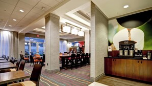 A hotel lobby with a long dining table, multiple chairs, a coffee station, and a large wall mural.